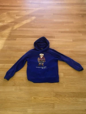 Polo Ralph Lauren ungdom storlek L 14-16 år - Säljer en blå hoodie från Polo Ralph Lauren med ett stort tryck av deras ikoniska björn på bröstet. Hoodien har huva och ribbade muddar vid ärmar och nederkant. Perfekt för dig som gillar streetwear och vill ha något bekvämt och snyggt.