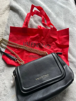 Svart handväska från Valentino - Snygg svart handväska från Valentino med guldfärgad kedjerem och lock med logga framtill. Väskan är äkta och är som ny. Levereras med röd dustbag med Valentino-tryck.