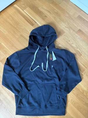 Polo Ralph Lauren Hoodie  - Snygg marinblå hoodie från Polo Ralph Lauren med klassisk huva och dragsnören. Har en liten broderad logga på bröstet och en stor magficka framtill. Tillverkad i mjukt material som känns skönt mot huden. Perfekt för en avslappnad och stilren look.