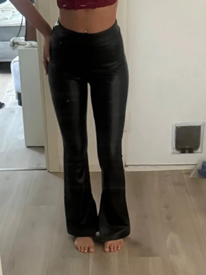 Svarta utsvängda leggings i skinnimitation - Säljer ett par svarta leggings i skinnimitation med hög midja och utsvängda ben. Sitter tight över höfterna och låren och har en snygg glansig finish. Säljs inte längre.