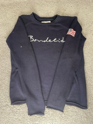 Marinblå BondeLid stickad tröja  - Mörkblå stickad tröja från BondeLid med vit broderad logga på bröstet och en klassisk BondeLid amerikansk flagga på ärmen. Tröjan har rund halsringning och långa ärmar. Materialet är stickat och ger en chill laidback look. Storlek = S