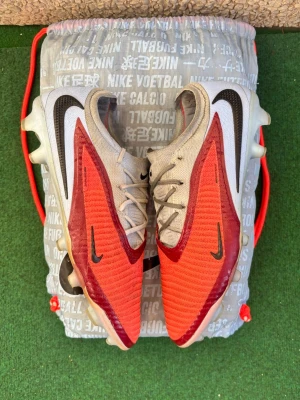 Nike röda och beige fotbollsskor - Snygga fotbollsskor från Nike i rött och beige med svarta detaljer och stora Swoosh-loggor på sidorna. Skorna har snörning och mönstrad ovansida för bättre bollkontroll. Yttersulan är genomskinlig med fasta dobbar, perfekt för spel på gräs. Vid bild 5 ser man att de bara en liten defekt men de gör inget de bara en liten öppning och man känner inte när man spelar. Box påse inkluderar✅ Går alltid att diskutera 