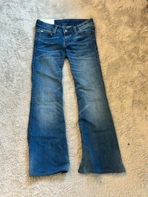 Blå bootcut jeans med låg midja - Snygga blå bootcut jeans med låg midja och klassiska fickor. Jeansen har slitningar framtill för en trendig look och stängs med knapp och dragkedja. Tillverkade i denim med lätt stretch för skön passform.