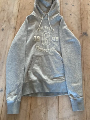 Grå hoodie med tryck och huva - Säljer en grå hoodie med vit text och siffror på bröstet: 'NEW HAVEN', '1949', 'CONNECTICUT' och ett stort G. Hoodien har huva med dragsko och en stor magficka. Tillverkad i mjukt sweatshirtmaterial och har ribbade muddar vid ärmar och nederkant.
