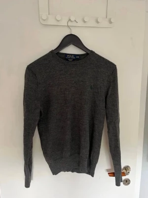 Mörkgrå stickad tröja från Polo Ralph Lauren - Mörkgrå stickad tröja från Polo Ralph Lauren med rund halsringning och diskret broderad logga på bröstet. Tröjan har långa ärmar och ribbade muddar vid ärmslut och nederkant. Perfekt för lager på lager och enkel att matcha. Storlek M men känns mer som S.