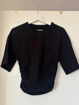 Svart topp med rynkade sidor - Svart topp med rund hals och korta ärmar. Toppen har snygga rynkade detaljer på sidorna som ger en cool och trendig look. Perfekt att styla med jeans eller kjol för en avslappnad vibe. Markerad midja.