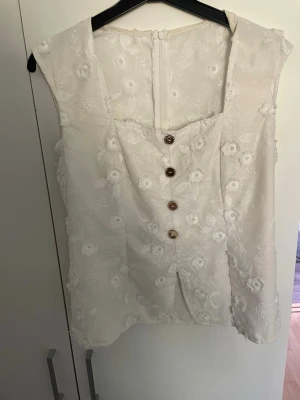 Vit blommig blus med knappar - Säljer en söt vit blus med broderade blommor och dekorativa knappar framtill. Blusen är ärmlös och har en lätt v-ringad halsringning. Perfekt för dig som gillar romantiska detaljer och en feminin look.