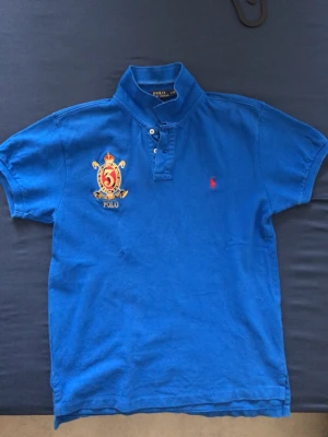 Polo Ralph Lauren - Blå piqué från Polo Ralph Lauren med röd logga och broderat emblem på bröstet.
