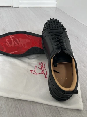 Svarta Louboutin sneakers med nitar - Snygga svarta sneakers från Christian Louboutin med ikoniska röda sulor och svarta nitar över tån. Skorna har en låg profil, snörning och är tillverkade i slätt läder med beige innerfoder. Perfekta för dig som vill sticka ut med en edgy och lyxig stil.