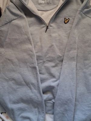 Ljusblå halvzip sweatshirt - Säljer en ljusblå sweatshirt med halv dragkedja och broderad gul logga på bröstet. Tröjan har en avslappnad passform och är tillverkad i mjukt bomullsmaterial. Perfekt för en chill och stilren look.