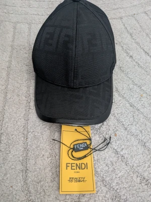 Svart keps från Fendi med FF-mönster - Snygg svart keps från Fendi med det klassiska FF-mönstret ton-i-ton över hela kepsen. Justerbar rem bak i läder och detaljerad med Fendi-logga på insidan. Tillverkad i ett slitstarkt tyg som känns exklusivt och trendigt.