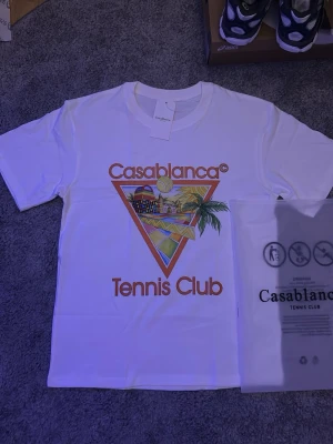 Vit Casablanca Tennis Club t-shirt - Säljer en vit t-shirt från Casablanca med färgglatt tryck och texten 'Tennis Club' framtill. Klassisk passform och rund hals. Perfekt för dig som gillar sportig och trendig stil.