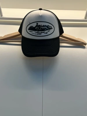 Svartvit truckerkeps med tryck corteiz  - Snygg svartvit truckerkeps med mesh baktill och justerbar snapback. Framsidan har ett svart tryck av ett landskap i oval ram. Klassisk böjd skärm och ventilerande material gör den perfekt för alla tillfällen.