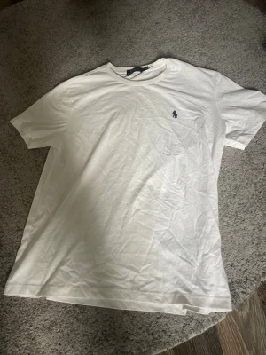 Vit t-shirt från Polo Ralph Lauren - Klassisk vit t-shirt från Polo Ralph Lauren med den ikoniska lilla loggan broderad på bröstet. T-shirten har rund hals och korta ärmar, tillverkad i mjuk bomull för en skön och avslappnad stil.