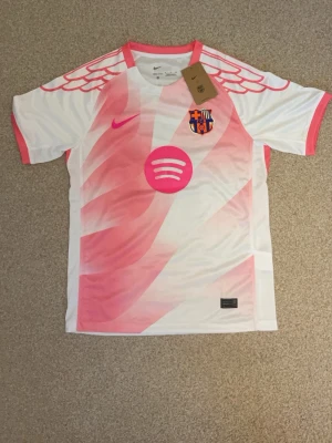 Barcelona Lamine Yamal matchtröja - Säljer en helt ny oanvänd vit och rosa FC Barcelona matchtröja från Nike med Lamine Yamal och nummer 10 på ryggen. Tröjan har rosa detaljer, klubbmärke, Spotify-logga och UNHCR-tryck. Tillverkad i lätt och ventilerande material, perfekt för fotboll eller träning.