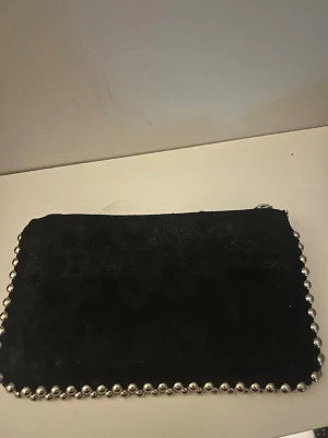 Svart kuvertväska med nitar från Zara - Snygg svart kuvertväska från Zara i mjukt material med silverfärgade nitar längs kanterna. Väskan har dragkedja upptill och en stilren, rektangulär form. Perfekt accessoar för att lyfta din outfit.