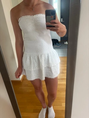 Vit axelbandslös kort klänning - Söt vit axelbandslös klänning med smock upptill och volangkjol i broderad bomull. Klänningen har en feminin och somrig look med två lager volanger och fina spetsdetaljer längst ner. Perfekt för varma dagar och studentfirande.