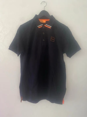 Svart Hugo boss pikétröja med orange detaljer - Snygg svarthugo boss  pikétröja med korta ärmar och orange samt vita detaljer på kragen. Tröjan har knappar framtill och en liten logotyp på bröstet. Perfekt till en avslappnad men stilren look.