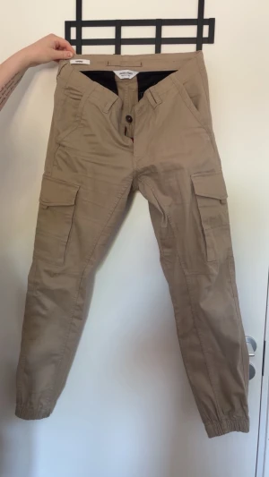 Beige cargobyxor med fickor - Snygga beige cargobyxor från Jack & Jones med flera praktiska fickor på sidorna och resår vid bensluten. Byxorna har en avslappnad passform och stängs med knapp och dragkedja. Perfekta för en casual och trendig stil.