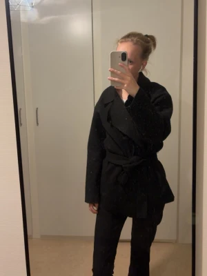 Svart kappa från Nelly  - Stilren svart kappa med bred krage och knytskärp i midjan. Kappan har en rak passform och är tillverkad i ett mjukt tyg som ger en snygg siluett. Perfekt för kyligare dagar och enkel att matcha med olika outfits.
