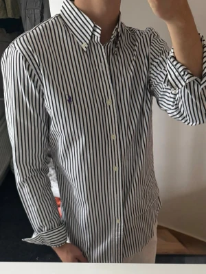 Randig skjorta med lång ärm - Snygg skjorta med svarta och vita vertikala ränder och klassisk krage. Skjortan har lång ärm, knappar framtill och en liten broderad logga på bröstet. Perfekt för dig som gillar stilrena plagg med en twist.