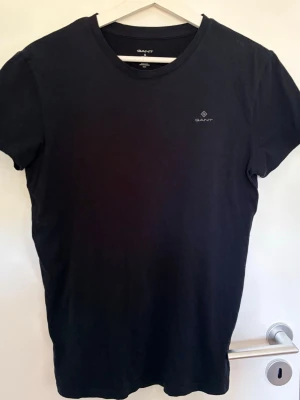 Svart t-shirt från GANT - Svart t-shirt från GANT med liten logga på bröstet. Klassisk rund halsringning och korta ärmar. Perfekt basic-plagg i garderoben, tillverkad i mjuk bomull för en skön känsla.