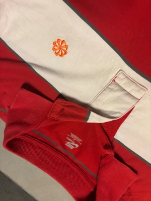 Randig pikétröja från Nike - Säljer en färgglad randig pikétröja från Nike med breda ränder i rött, vitt och turkost. Tröjan har klassisk krage, knappslå och en liten orange broderad blomma på bröstet. Perfekt för dig som gillar sportig och avslappnad stil.