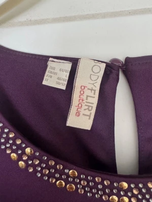 Lila ärmlös klänning Bodyflirt - 44/46 - Snygg lila ärmlös klänning från Bodyflirt med dekorativa guldfärgade och silvriga nitar vid halsringningen. Klänningen har en rak passform och knäpps med en knapp i nacken. Perfekt för dig som vill sticka ut lite extra.