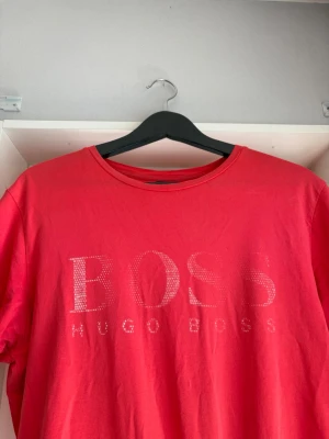 Röd kortärmad t-shirt från Hugo Boss - Snygg röd t-shirt från Hugo Boss med rund halsringning och stort BOSS-tryck framtill i vita prickar. Tillverkad i mjuk bomull som känns skön mot huden. Perfekt för dig som vill ha en enkel men ändå stilren look.