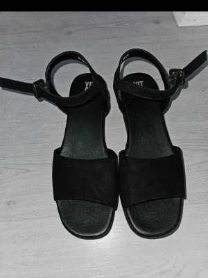 Svarta kilklackade sandaler - Stilrena svarta sandaler med kilklack och bred rem över tårna. Sandalerna har justerbart ankelband med spänne och är tillverkade i mockaimitation. Perfekta för dig som vill ha både höjd och komfort i en klassisk design. Använda en gång 