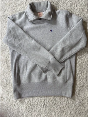 Grå halvzip hoodie från Champion - Snygg grå halvzip hoodie från Champion med liten broderad logga på bröstet. Tillverkad i mjuk bomullsblandning och har ribbade muddar vid ärmslut och nederkant. Perfekt för en chill och sportig look.