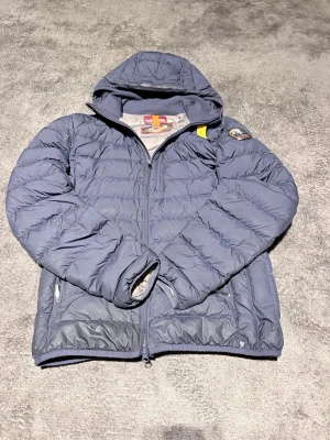 Blå dunjacka med huva Parajumpers - Snygg blå dunjacka från Parajumpers med huva och dragkedja framtill. Jackan har två sidofickor med dragkedja och en patch på ärmen. Insidan är ljusgrå och jackan är lätt vadderad för extra värme. Perfekt för kalla vinterdagar.