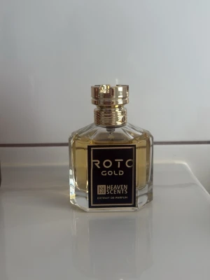 ROTO GOLD parfym från Heaven Scents - Exklusiv parfymflaska med guldigt lock och etikett. Flaskan är sexkantig i klart glas och innehåller en gyllene vätska. Märkt med 'ROTO GOLD' och 'Heaven Scents Extrait de Parfum' på framsidan. Perfekt för dig som gillar lyxiga dofter.