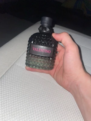 Valentino Uomo parfym - Snygg parfymflaska från Valentino med svart och mörkgrön glasdesign och nitar över hela flaskan. Flaskan har en svart kork och en etikett med rosa text. Perfekt för dig som gillar exklusiva och trendiga dofter.