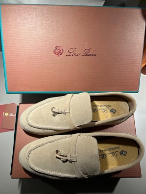 Beige mocka loafers från Loro Piana - Snygga beige loafers i mocka från Loro Piana med klassisk design och dekorativ metall-detalj framtill. Skorna har en stilren siluett och mjuk ovandel i mocka som ger en exklusiv känsla. Perfekta för dig som gillar tidlös och elegant stil.