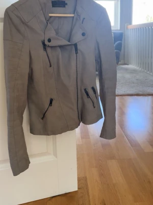 Beige skinnjacka med dragkedjor från only  - Snygg beige skinnjacka med flera svarta dragkedjor och cool asymmetrisk krage. Jackan har figurnära passform och detaljerade sömmar på ärmarna för en modern look. Perfekt för dig som vill ha en stilren och trendig jacka.