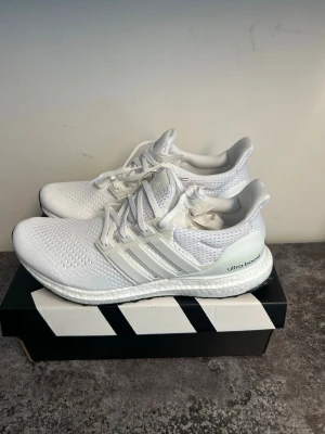 Vita Adidas Ultra Boost sneakers - Fräscha helvita Adidas Ultra Boost sneakers med stickad ovandel och klassiska tre ränder på sidan. Skorna har en bekväm Boost-sula och ventilerande meshmaterial för extra komfort. Perfekta för dig som gillar stilrena och sportiga sneakers.