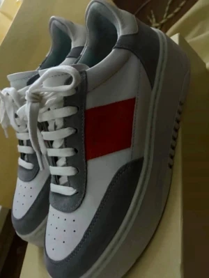 Axel Arigato sneakers med orange detalj - Snygga sneakers från Axel Arigato i vitt och grått med en tydlig orange detalj på sidan. Skorna har vita snören, perforerad tå och grå mockadetaljer. Klassisk låg modell med chunky sula och stilren design som sticker ut.