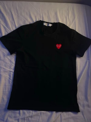 CDG T-Shirt - Svart & rött hjärta - storlek S - Svart t-shirt med ett ikoniskt rött hjärta med ögon broderat på bröstet. Klassisk rund hals och korta ärmar. Tillverkad i mjuk bomull för en skön känsla. Perfekt för dig som gillar stilrena plagg med en twist. Använd 1 gång.