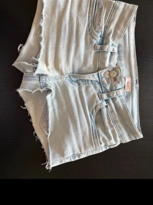 Ljusblå jeansshorts  - Säljer ett par ljusblå jeansshorts och dubbla knappar i midjan. Shortsen har fem fickor och rosa broderade detaljer på bakfickorna. Perfekta för varma dagar och en avslappnad stil.