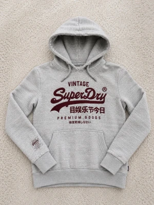 Grå Superdry hoodie med tryck - Snygg grå hoodie från Superdry med stor vinröd logga och japansk text på bröstet. Klassisk känguruficka framtill, dragsko i huvan och ribbade muddar. Perfekt för en avslappnad streetwear-look.
