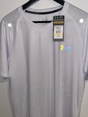 Vit tränings-tshirt från Under Armour - Säljer en vit tränings-tshirt från Under Armour med reflekterande logga på bröstet och ryggen. Tillverkad i ett lätt och snabbtorkande funktionsmaterial som andas, perfekt för löpning eller gym. Klassisk passform med rund hals och korta ärmar.