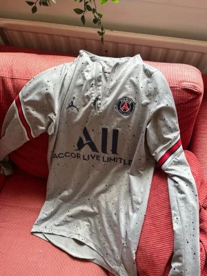 Grå PSG långärmad träningströja/ behöver lagas lite  - Snygg grå långärmad träningströja från PSG x Jordan med svartprickigt mönster och dragkedja vid halsen som tyvärr gått av… kan lagas… Tröjan har PSG-logga på bröstet, svarta och röda detaljer på ärmarna samt tryck framtill. Perfekt för fotbollsträning eller gymmet.