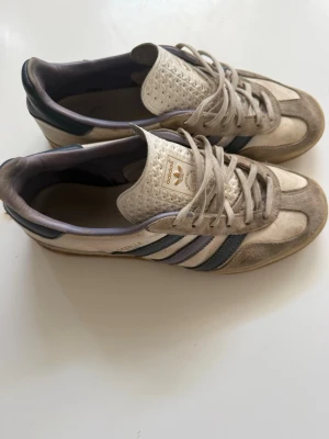 Adidas Gazelle beige sneakers - Snygga Adidas Gazelle sneakers i beige mocka med klassiska mörkblå ränder på sidan och gulddetaljer. Skorna har låg profil, vit plös med struktur och gummisula. Perfekta för en avslappnad och trendig stil.