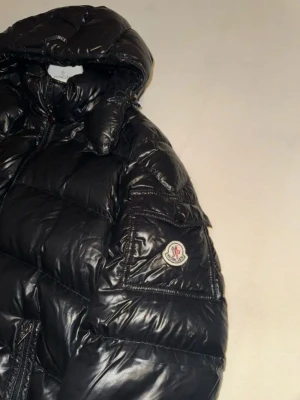 Svart dunjacka från Moncler - Snygg svart dunjacka från Moncler med glansig finish och klassisk pufferdesign. Jackan har huva, dragkedja och Moncler-logga på ärmen. Perfekt för kalla vinterdagar och riktigt varm tack vare dunfyllningen.
