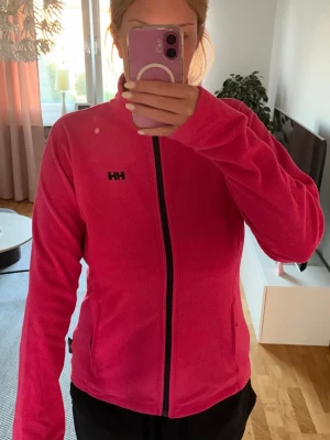 Rosa fleecejacka från Helly Hansen - Rosa fleecejacka från Helly Hansen med hel dragkedja framtill och två fickor med dragkedja. Jackan har hög krage och är tillverkad i mjukt fleecematerial.