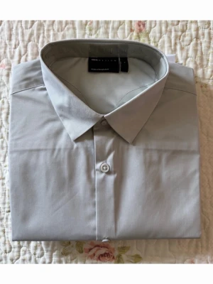 Ljusgrå skjorta från ASOS DESIGN - Säljer en stilren ljusgrå skjorta från ASOS DESIGN. Skjortan har klassisk krage, knäppning framtill och är tillverkad i ett mjukt bomullsmaterial. Perfekt för dig som gillar en clean och enkel look till sommaren. 