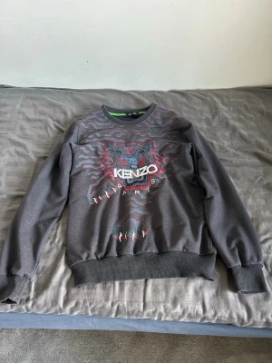 Grå sweatshirt från Kenzo med tiger - Säljer en grå sweatshirt från Kenzo med det ikoniska tigerhuvudet broderat framtill i blått, rosa och vitt. Tröjan har rund halsringning, ribbade muddar och nederkant. Materialet är mjukt och bekvämt, perfekt för en trendig streetwear-look.