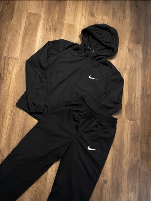 Svart Nike träningsset med huva - Säljer ett svart träningsset från Nike med matchande jacka och byxor. Jackan har huva och dragkedja, medan byxorna har resår i midjan och justerbar dragsko. Båda plaggen har den klassiska vita Nike-loggan. Perfekt för träning eller chill. Tyvärr vid armen är den lite sönder men inget man lägger märket till en kap om du frågar mig me detta priset. Skriv om du är intresserad eller har några föderningar.priset kan diskretas  