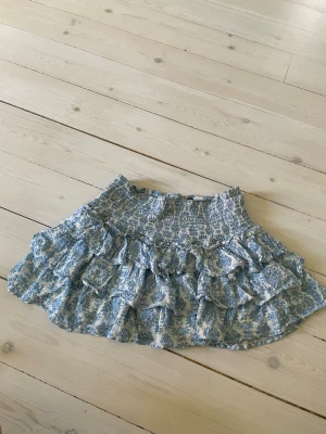 Blåvit volangkjol från Divided/HM - Supersöt kort volangkjol från Divided i blått och vitt med blommigt mönster. Kjolen har smock i midjan för skön passform och flera lager volanger som ger en lekfull look. Perfekt för varma dagar och somriga outfits(finns shorts under).